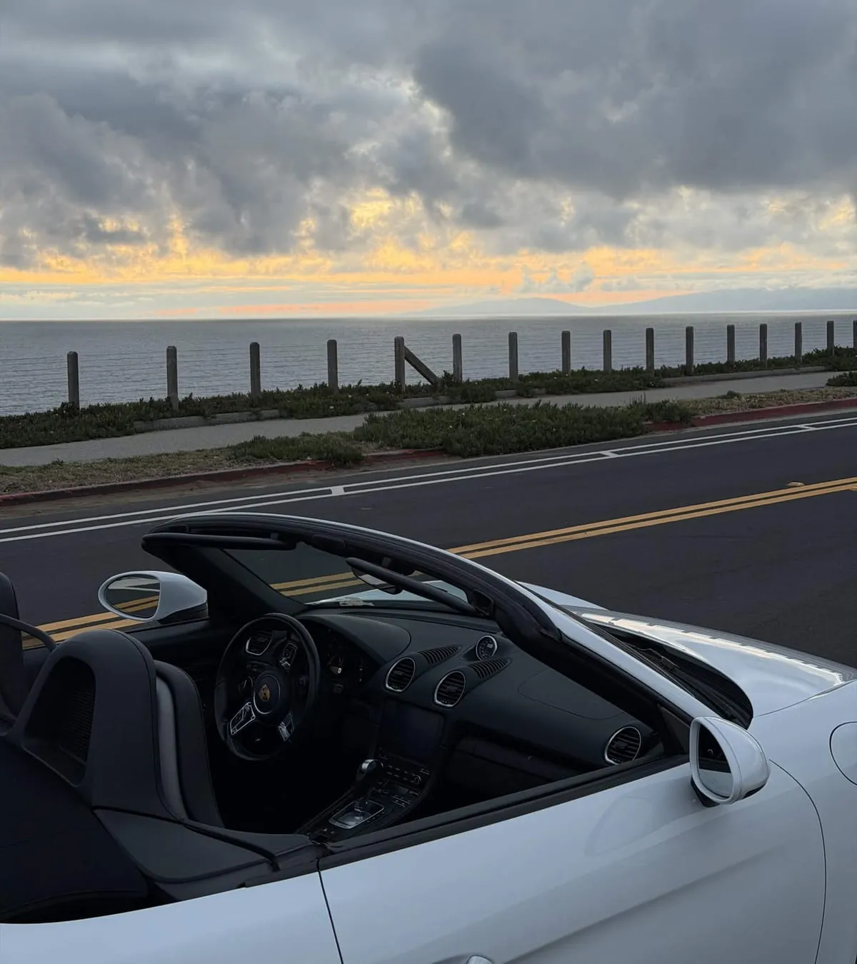 A white Porsche convertible parked on a coastal road at sunset, ocean and clouded sky beyond the guardrail.